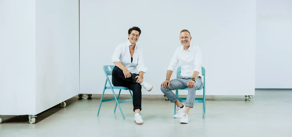 Die Coaches Stefanie Schmaus und Steffen Karpstein sitzen auf Klappstühlen in einer hellen Umgebung