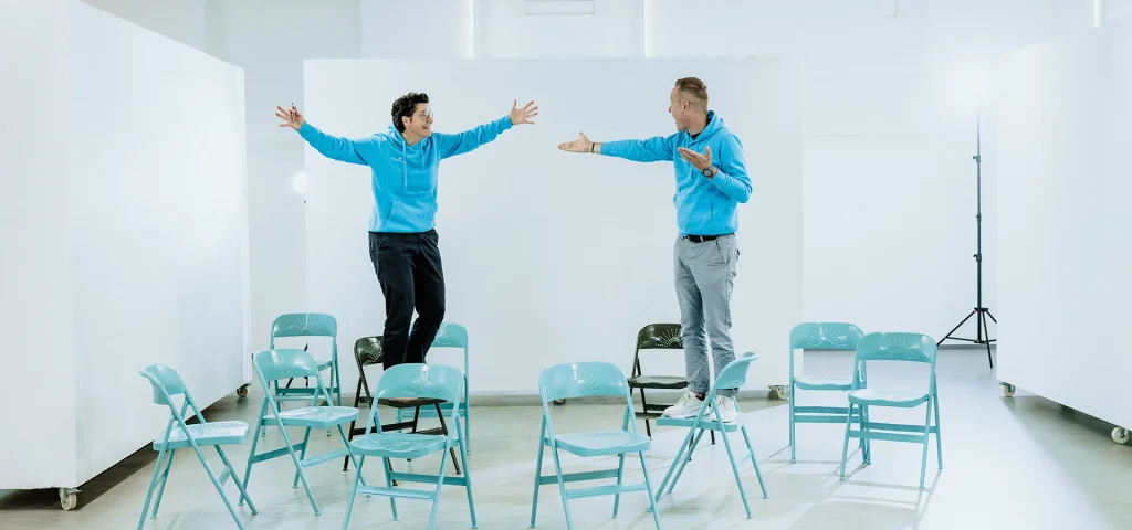 Die Coaches Stefanie Schmaus und Steffen Karpstein stehen auf Klappstühlen in einer hellen Umgebung